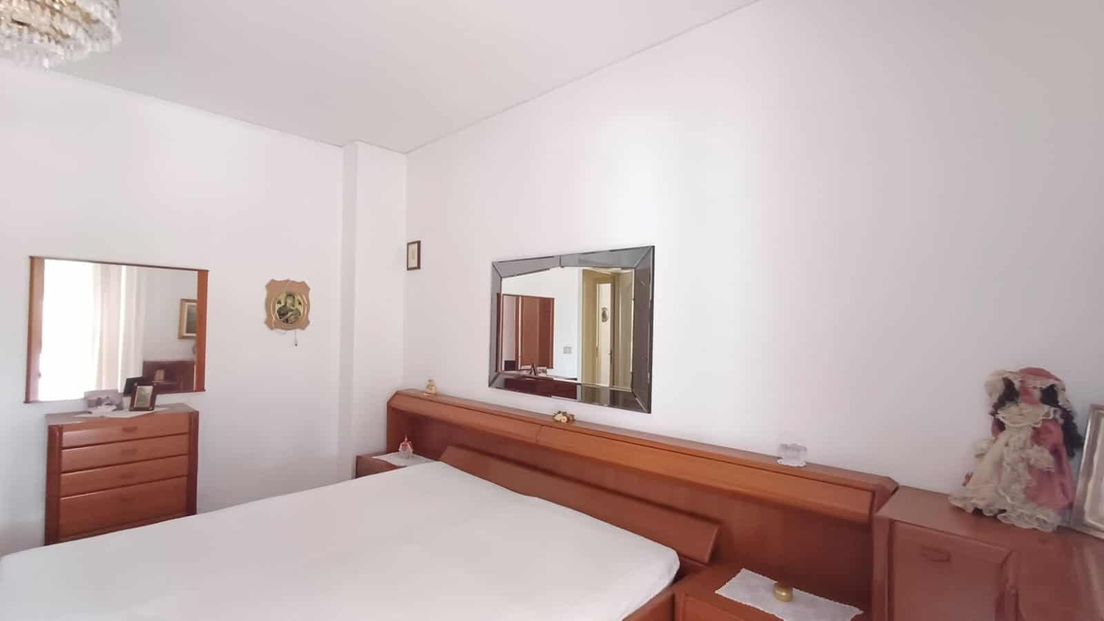Camera da letto luminosa con pareti bianche, grande specchio e letto matrimoniale in legno.