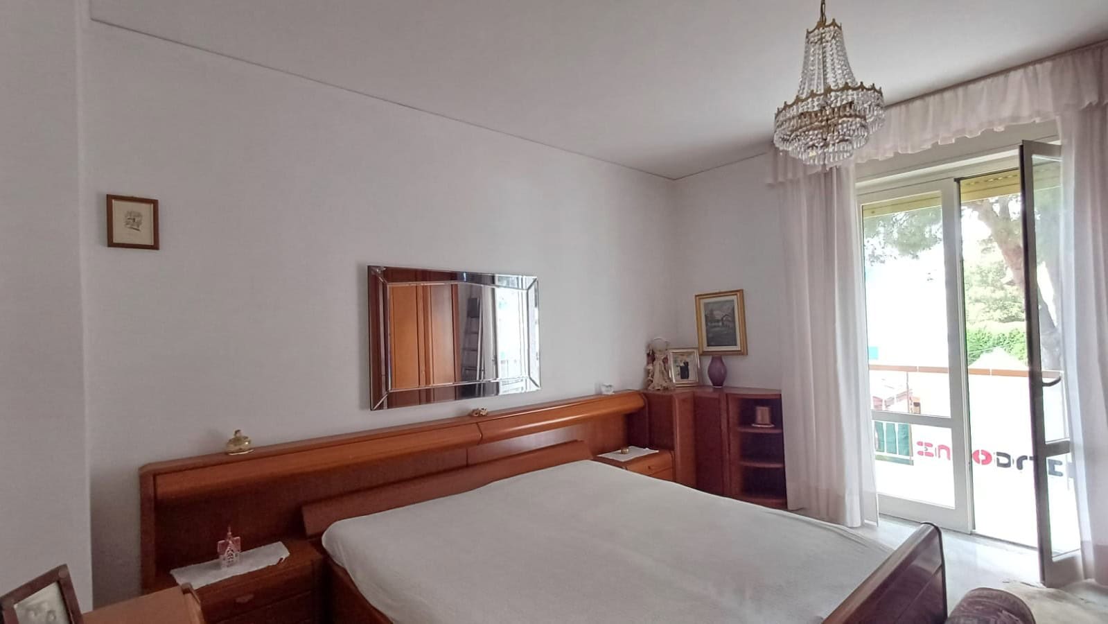 Camera che mostra una camera da letto luminosa con letto matrimoniale, armadio in legno, specchio e balcone con vista sul verde.
