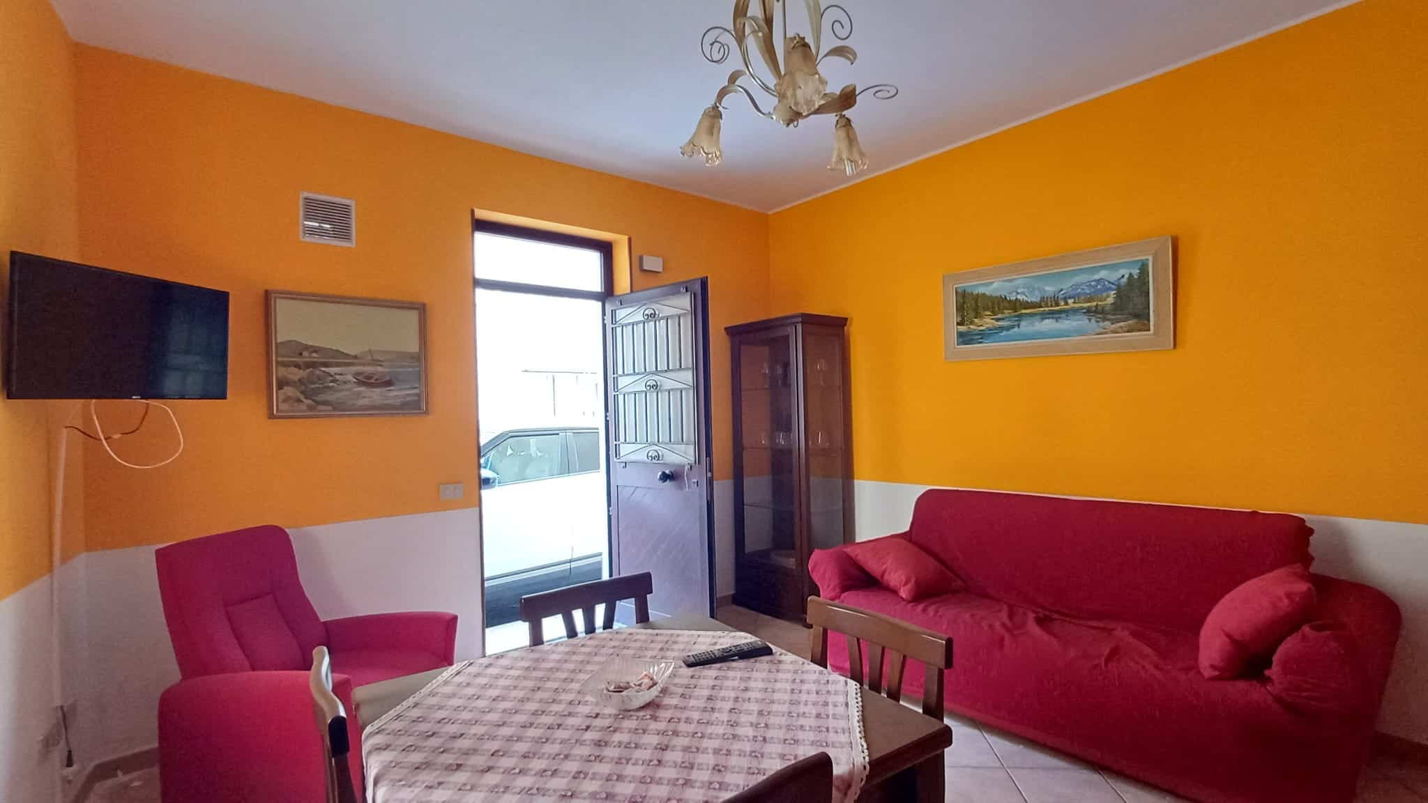 Interno di un appartamento con pareti arancioni, divano rosso e tavolo con sedie, vista sulla porta d'ingresso e decorazioni alle pareti.