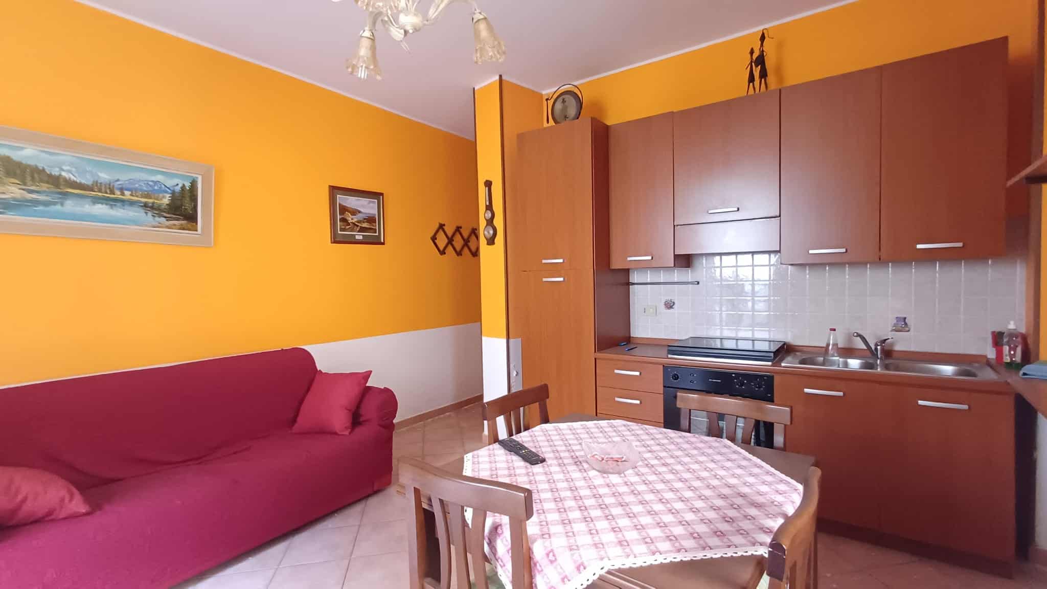 WhatsApp Image 2026-07-25 at 12.36.15dcfsdf Interno di un appartamento con pareti arancioni, divano rosso e cucina in stile semplice con mobili in legno e pareti rivestite di piastrelle.
