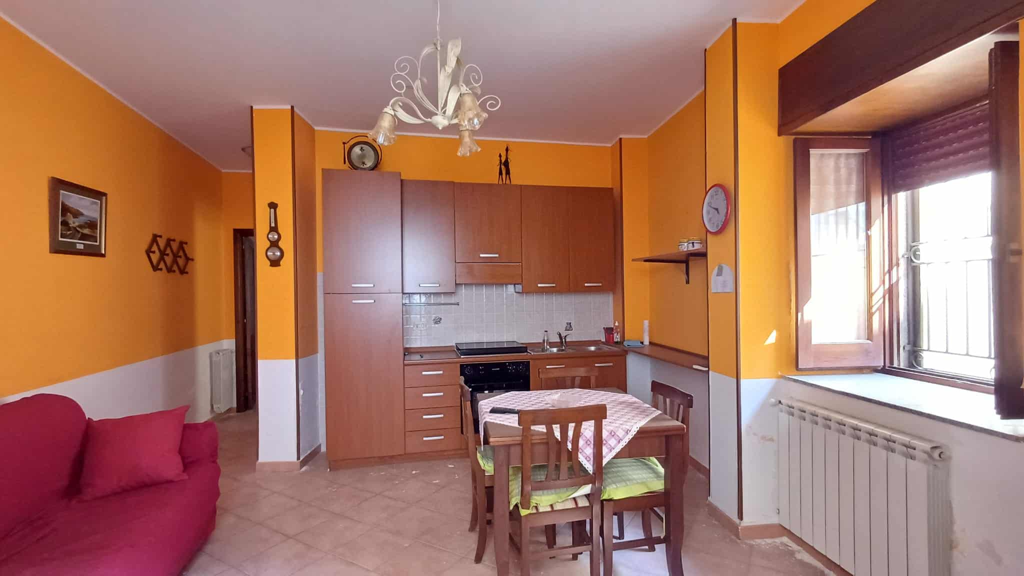 Interno di un appartamento con pareti arancioni, cucina moderna e zona soggiorno con divano rosso, finestra grande con vista sulla strada.