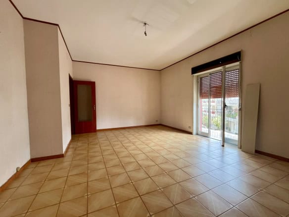 Ampio soggiorno vuoto con grandi finestre e accesso al balcone, pavimento in piastrelle beige, ideale per appartamento in vendita a Pace del Mela.