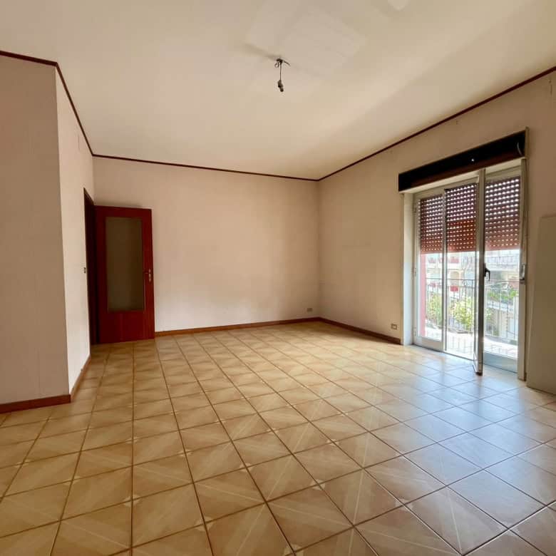 Ampio soggiorno vuoto con grandi finestre e accesso al balcone, pavimento in piastrelle beige, ideale per appartamento in vendita a Pace del Mela.