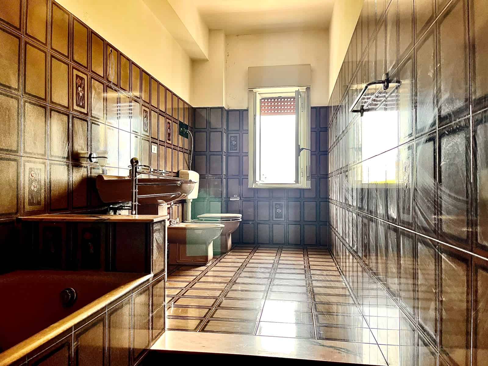 Bagno spazioso con pareti e pavimenti in piastrelle marroni, finestra luminosa e sanitari moderni in un appartamento in vendita a Pace del Mela.