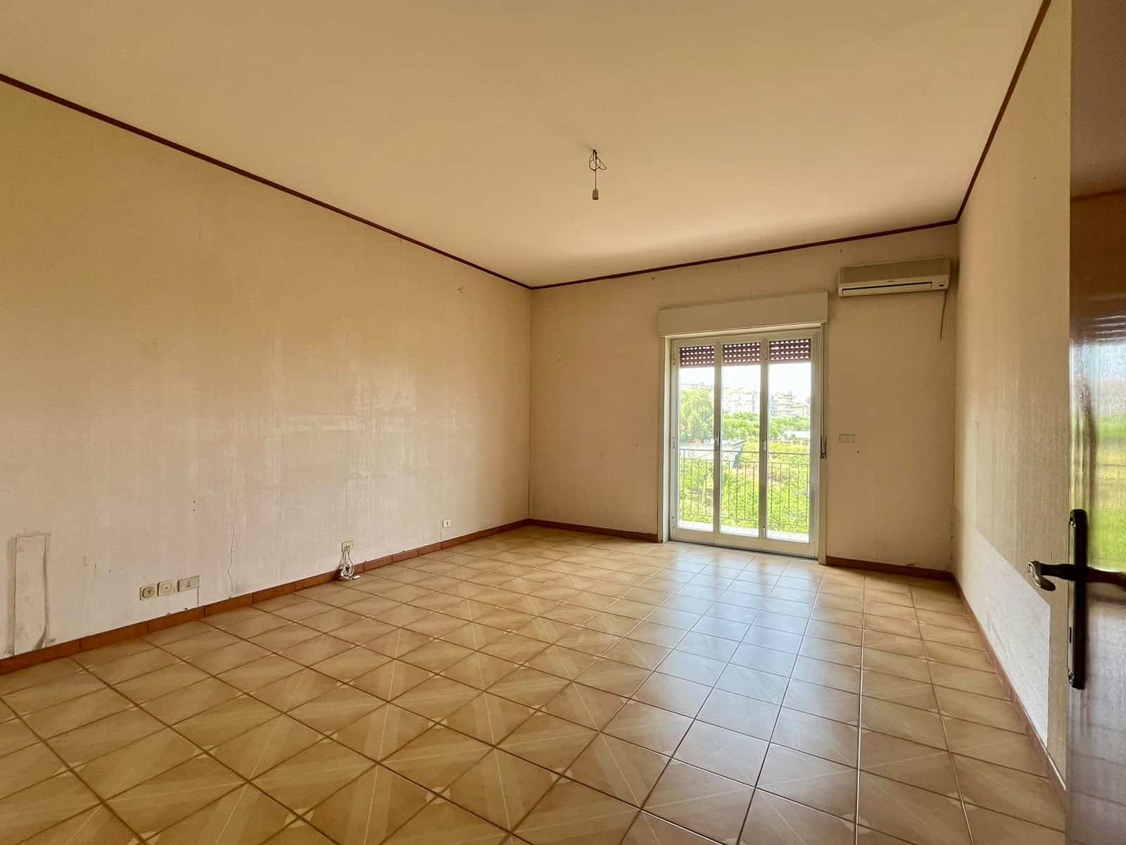 Ampio soggiorno vuoto con pareti beige e pavimento in piastrelle di ceramica, balcone con vista sul verde.