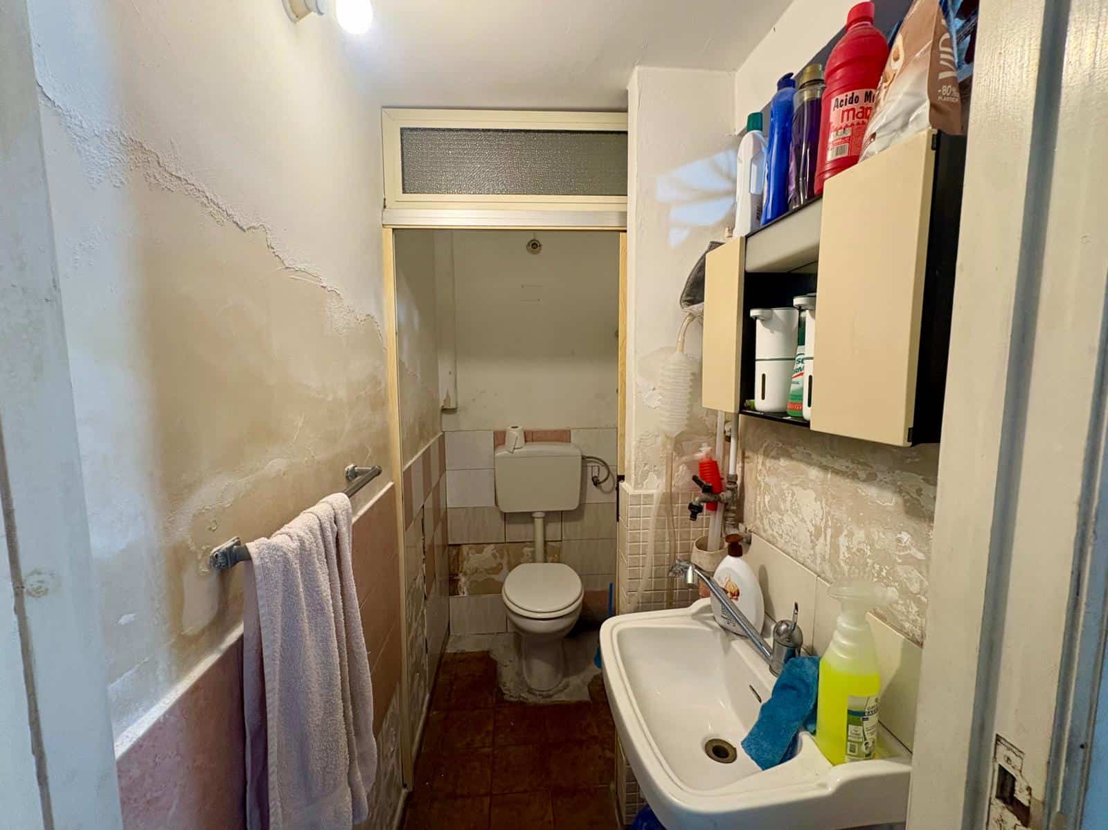 Bagno in condizioni di ristrutturazione con lavabo, WC e scaffali con prodotti di pulizia.