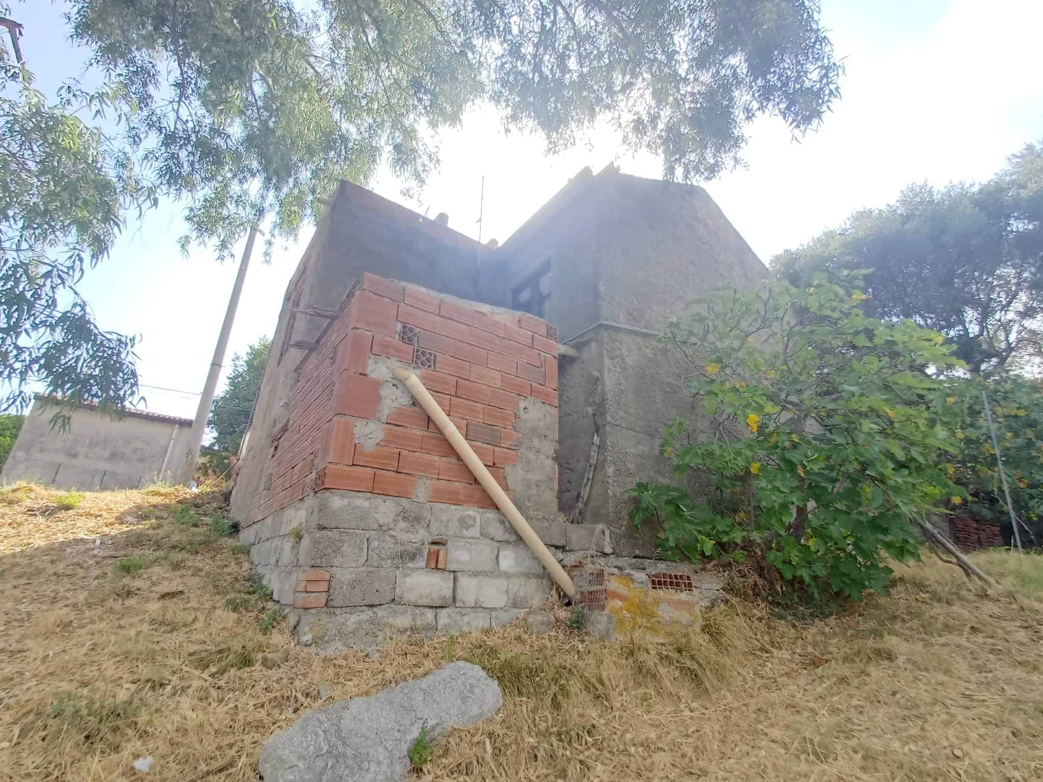 Struttura in costruzione con mattoni rossi e cemento, su un terreno in pendenza con alberi e vegetazione circostante.