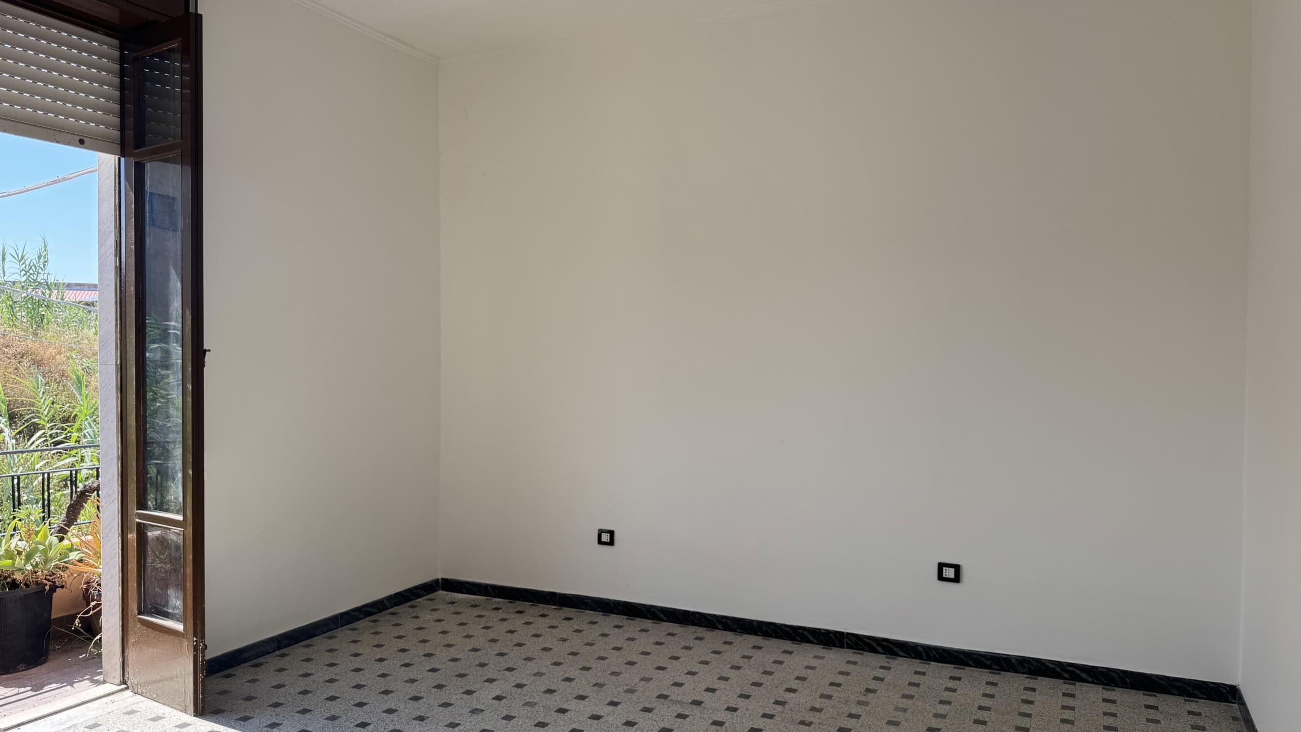 Stanza vuota con pareti bianche, pavimento in piastrelle e porta finestra che dà su un balcone con vista sul giardino.