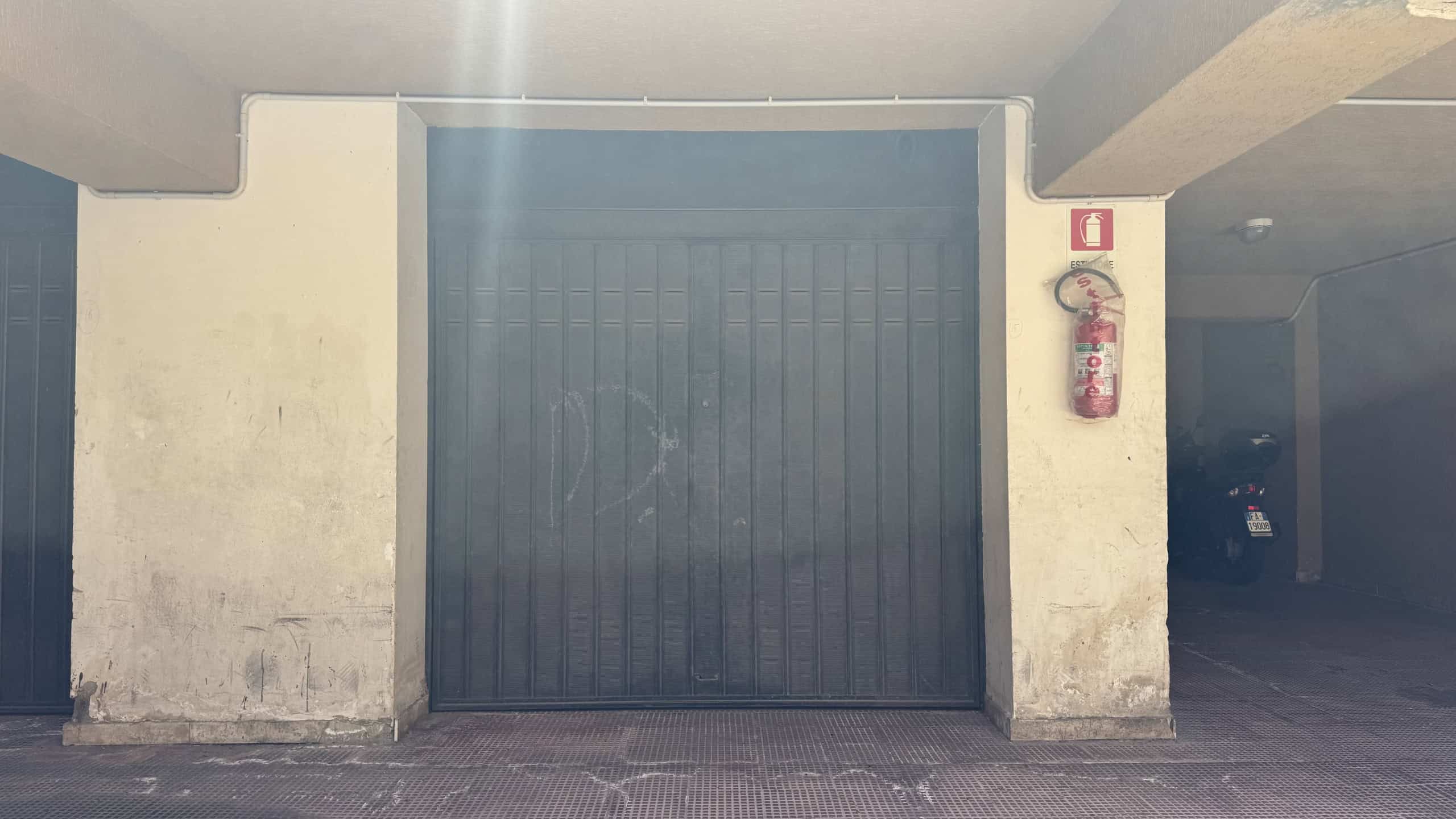 Portone di garage in un edificio di recente costruzione a Messina, con pareti in cemento e un estintore vicino.