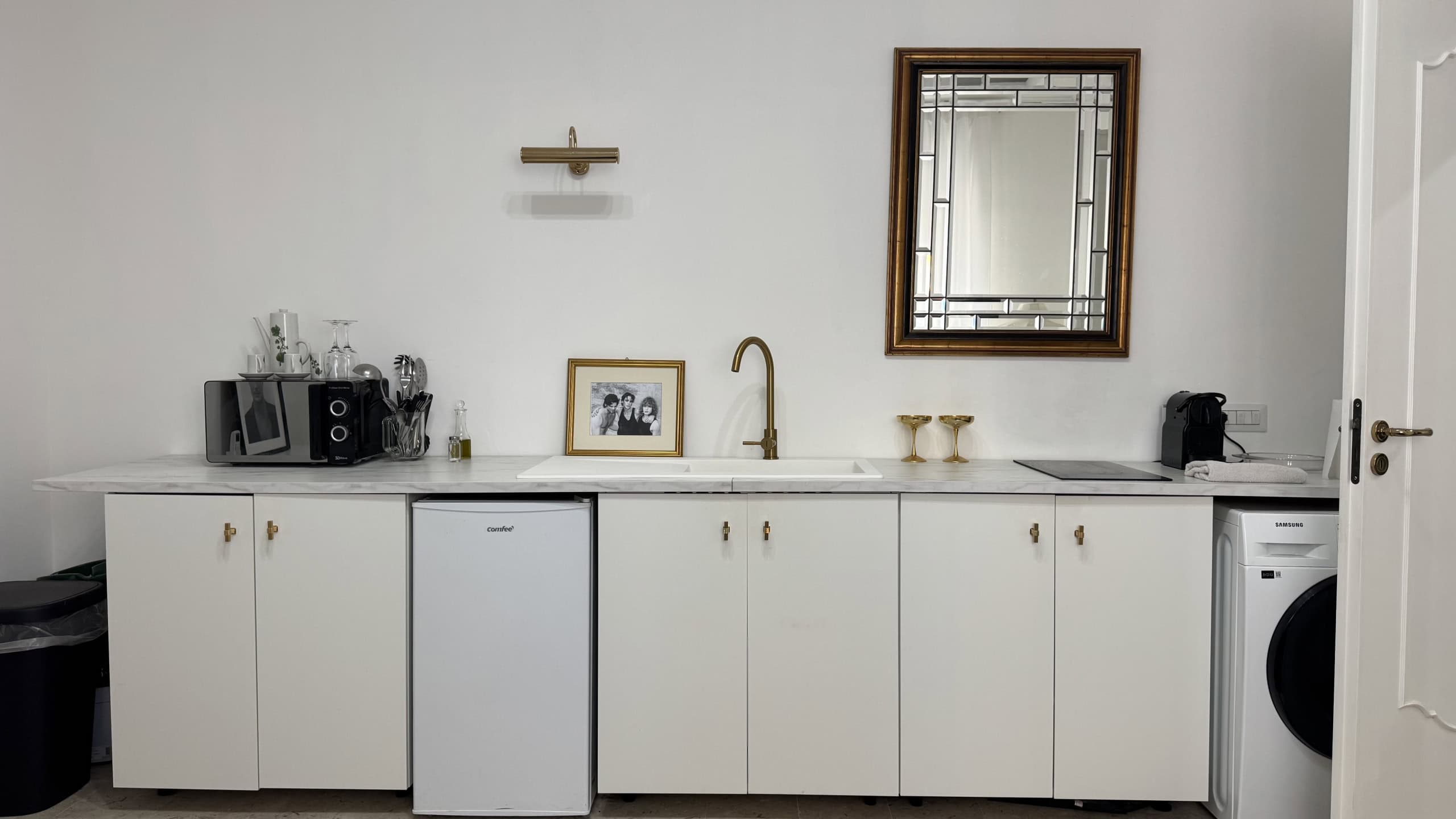 Angolo cucina moderna con mobili bianchi, lavabo, frigorifero e lavatrice, decorato con specchio e accessori minimalisti.