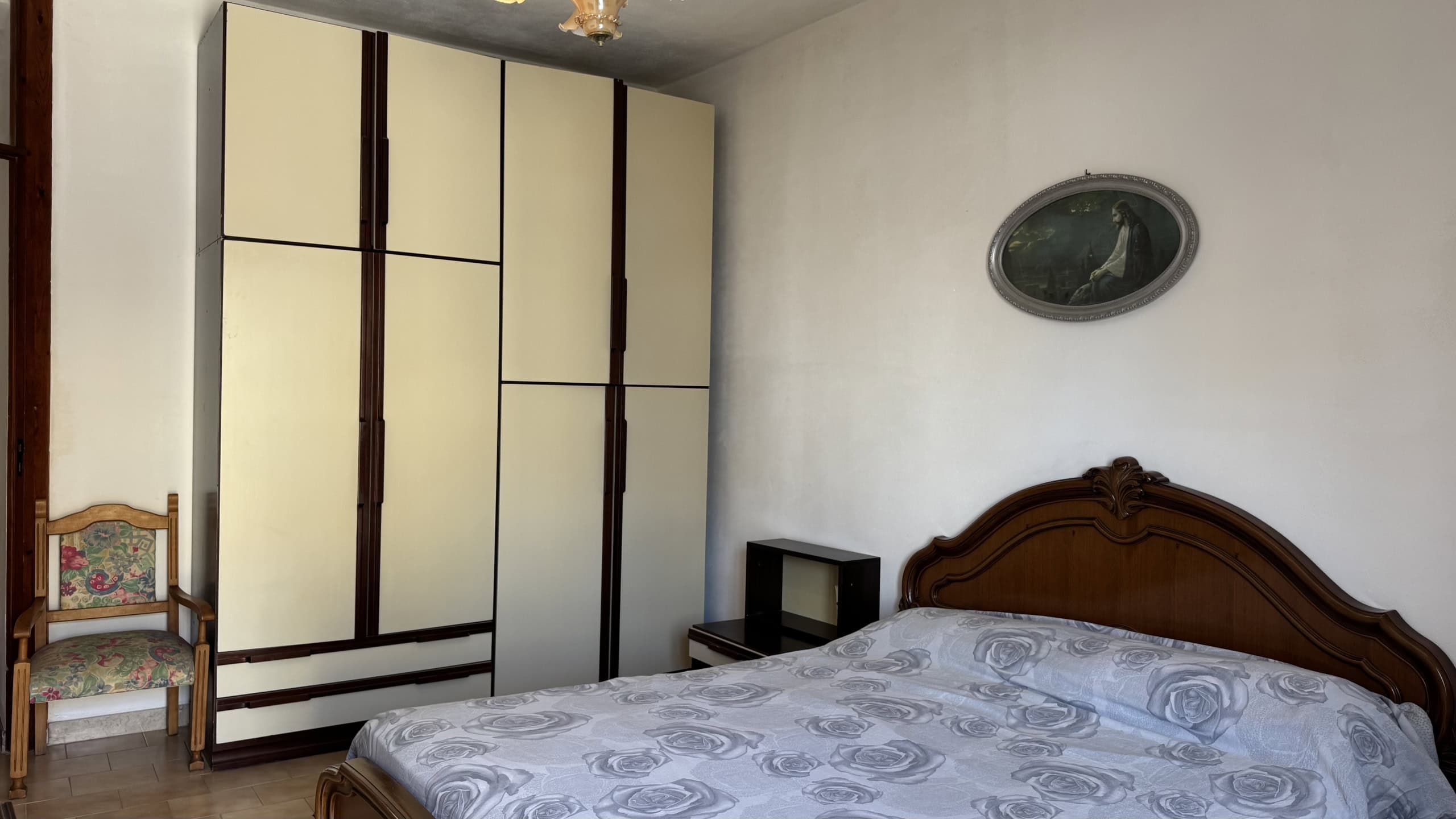 Camera da letto con armadio grande, letto in legno e sedia antica, pareti bianche e decorazione ovale sopra il letto.