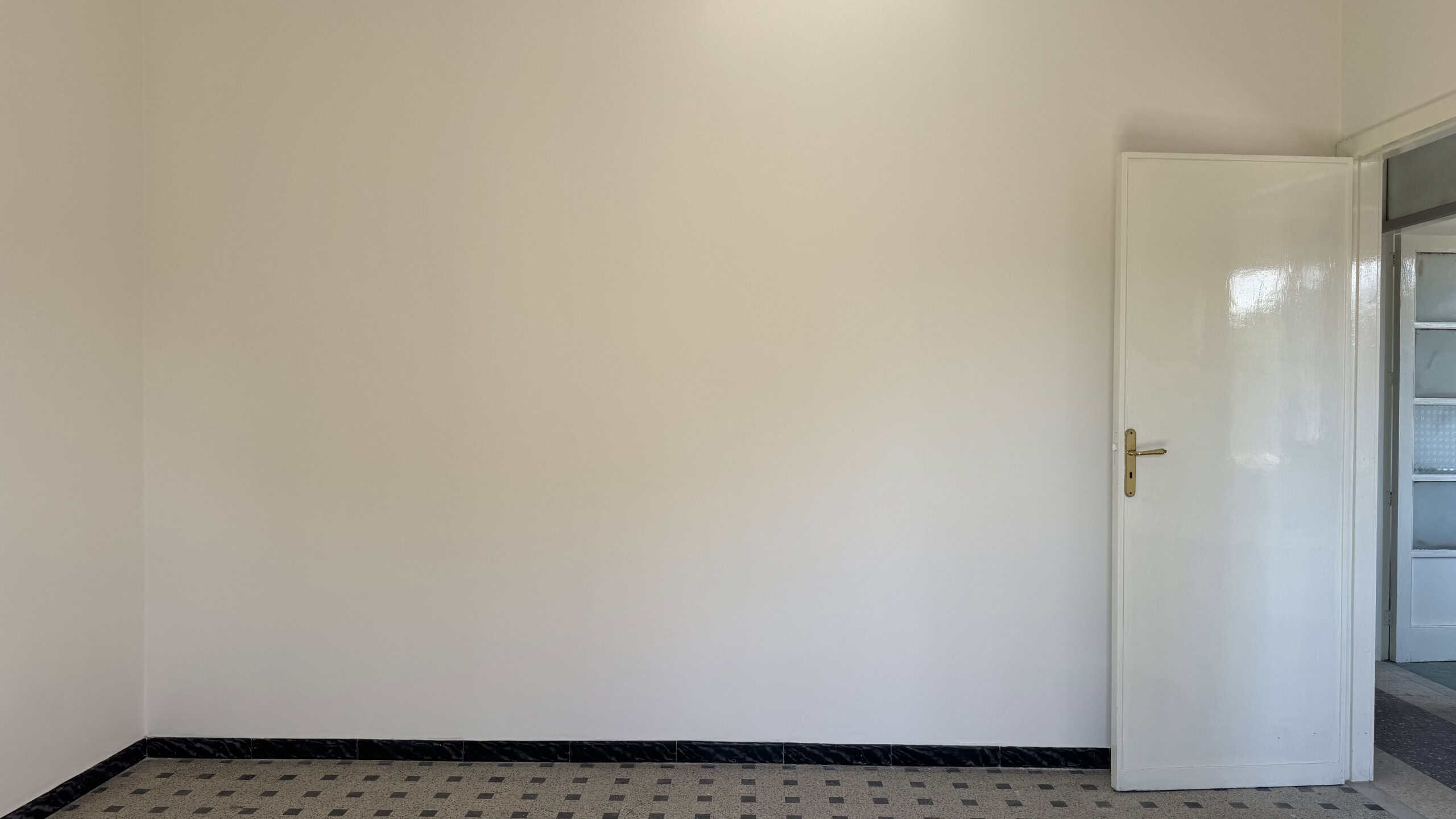 Interno di una stanza vuota con pareti bianche e pavimento in piastrelle nere, porta aperta che conduce a un balcone o terrazza.