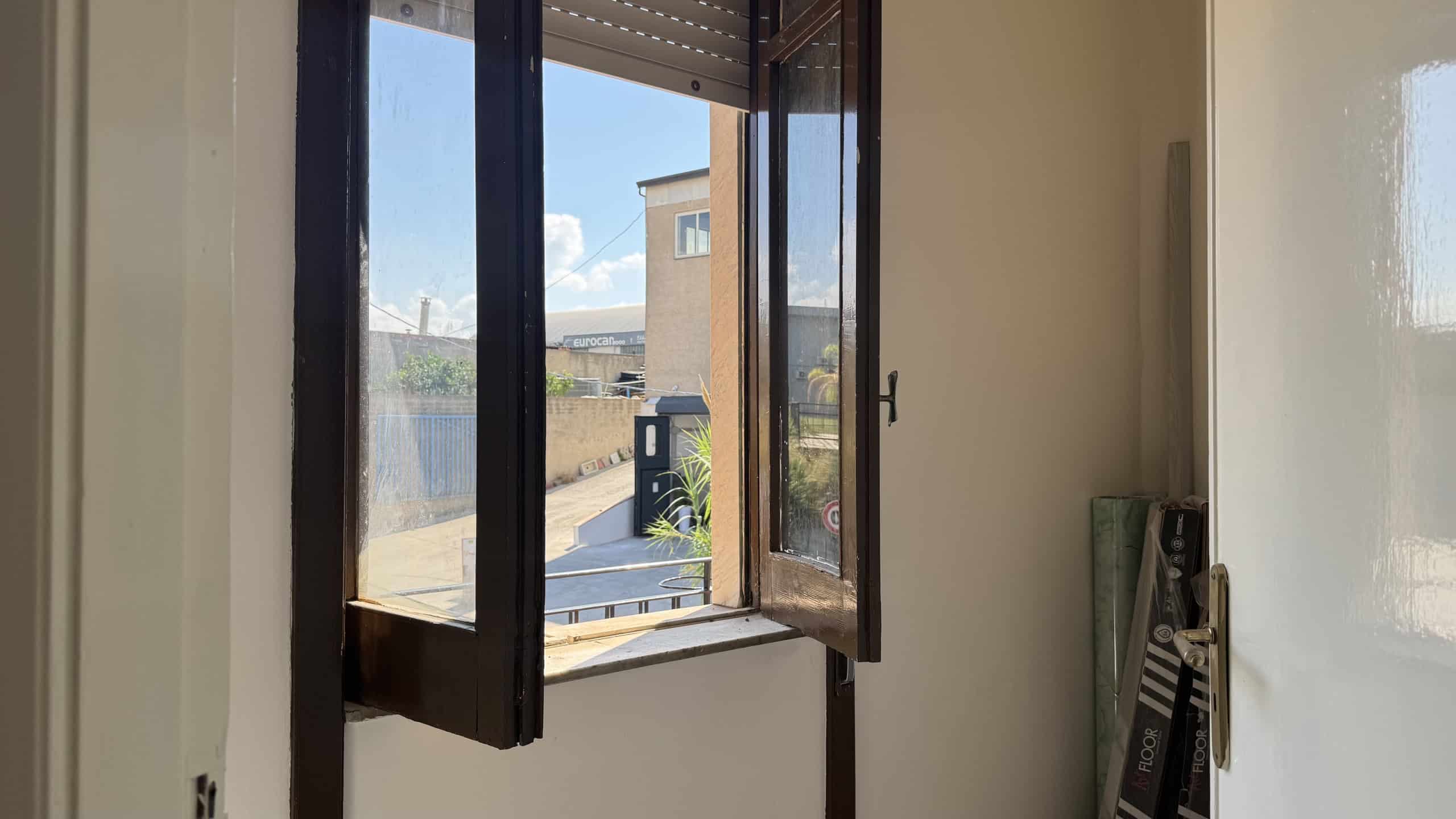 Finestra aperta in una stanza che mostra un cortile esterno con edifici e cielo blu.