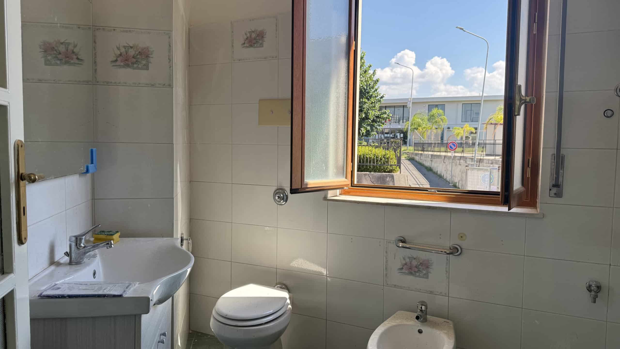 Bagno con finestra aperta, lavabo e WC in una casa in vendita a Venetico, Me.