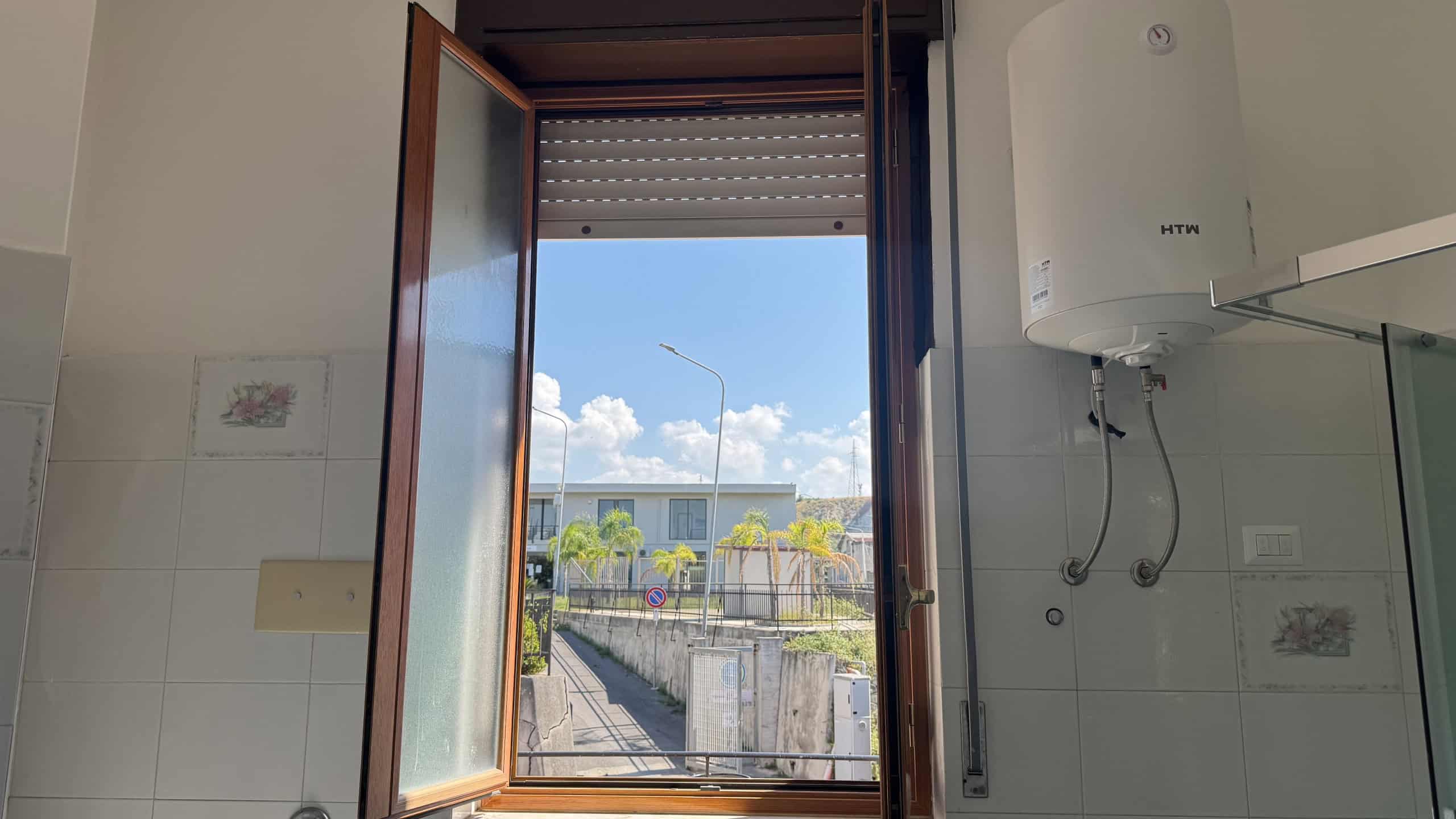 Finestra aperta che mostra una strada e il cielo azzurro con nuvole bianche, all'interno di una cucina moderna.