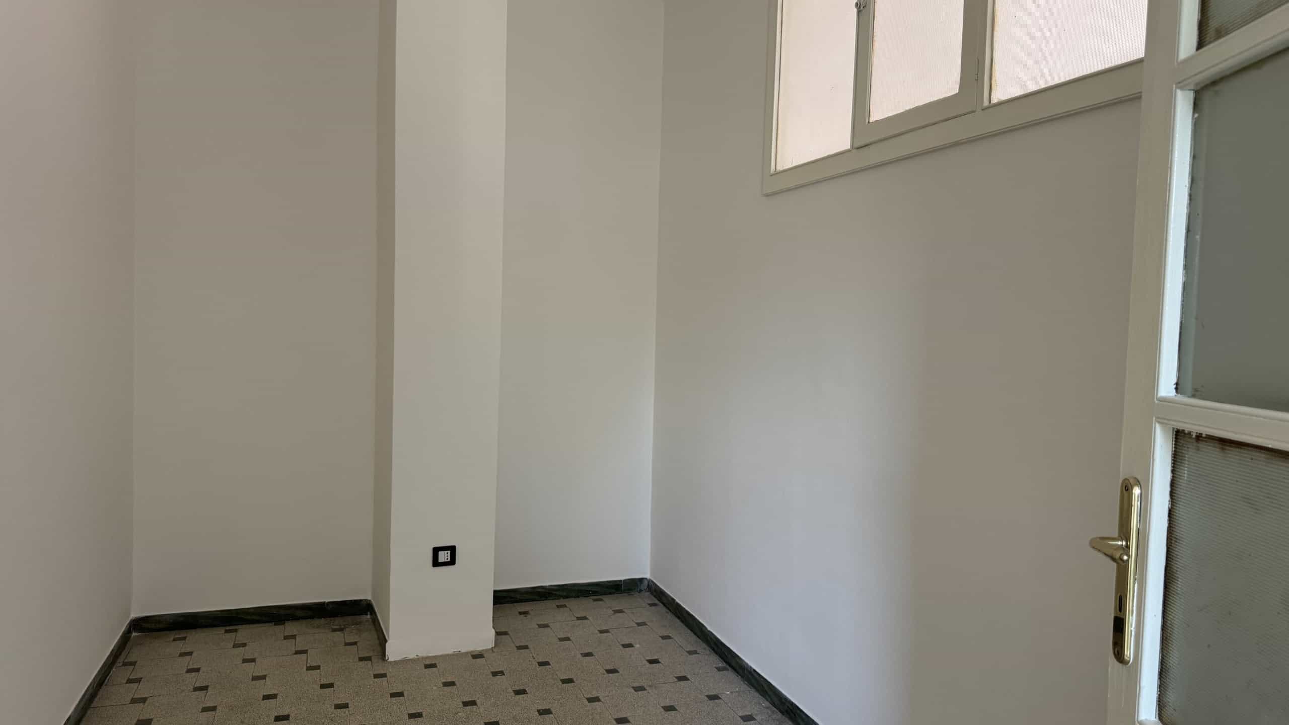 Interno di una stanza vuota con pareti bianche e pavimento in piastrelle beige e nere.