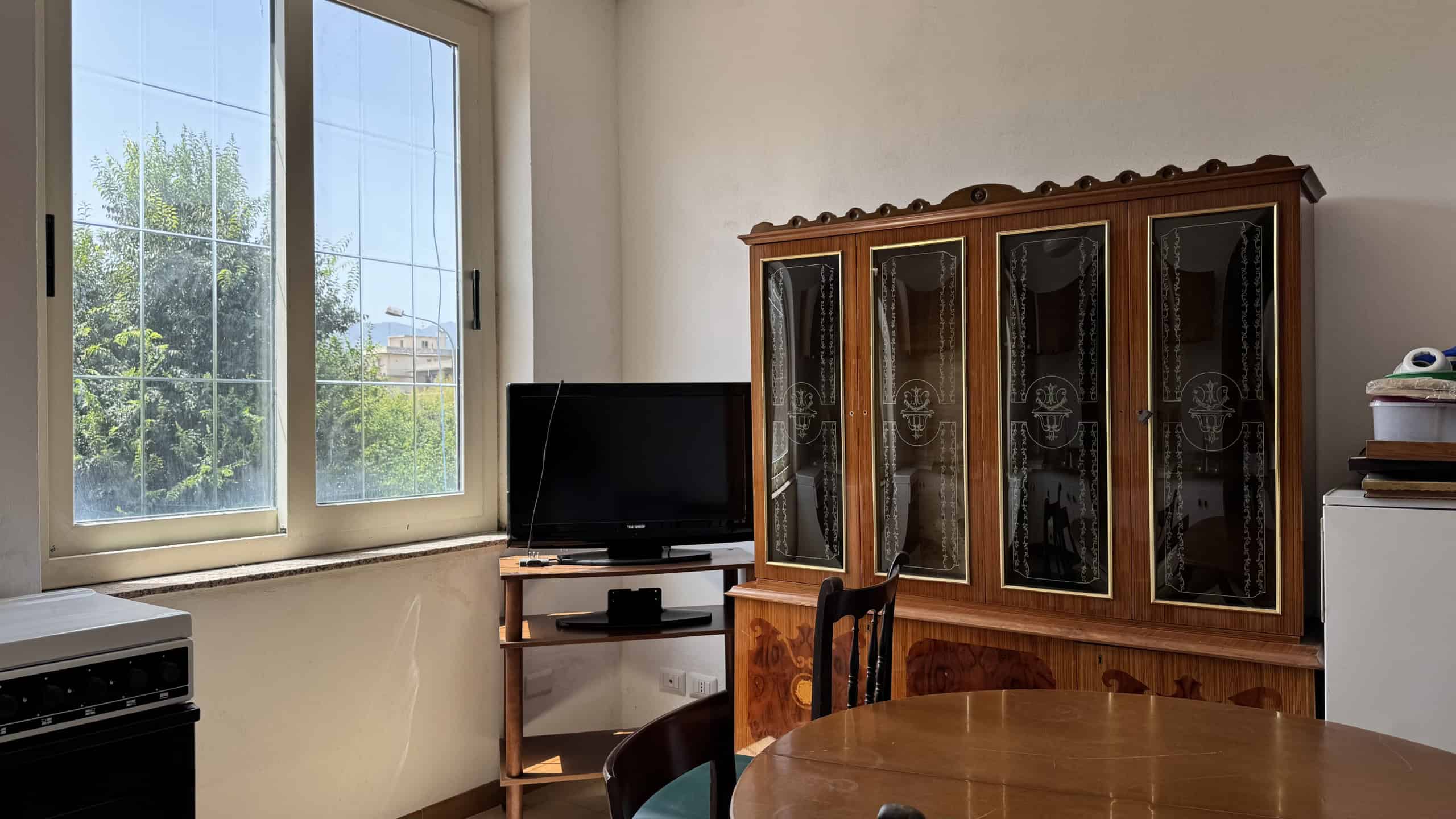 Interno di una casa con finestra, mobile in legno con vetrine e tavolo in legno, ambiente luminoso e accogliente.
