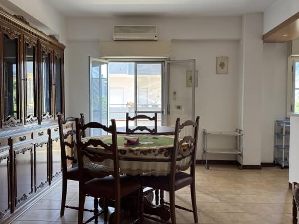Interno di un appartamento in vendita con sala da pranzo, tavolo rotondo e sedie in legno, pareti bianche e grande finestra con vista esterna.