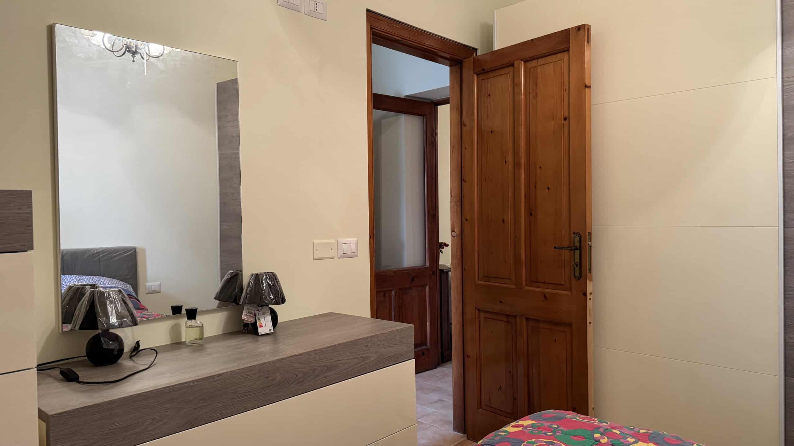Interno di una camera da letto con porta in legno, specchio e mobili moderni, in stile semplice e funzionale.