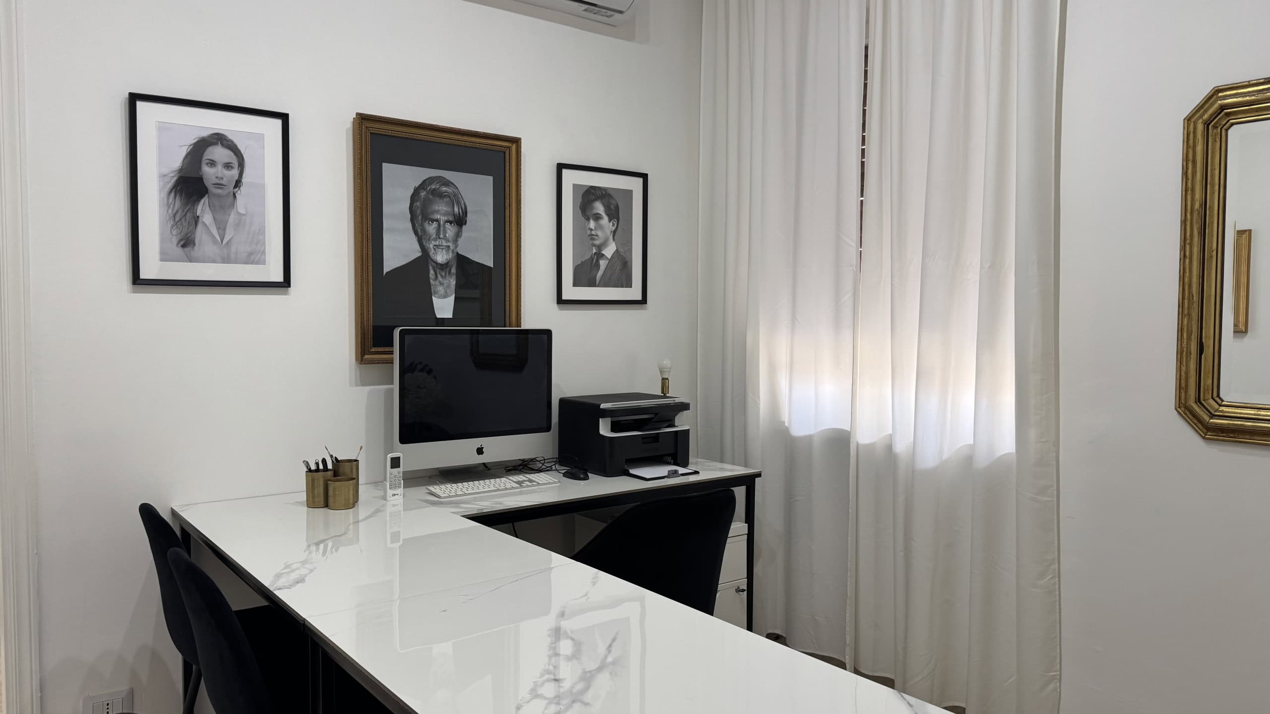 Ufficio moderno con scrivania bianca, computer e quadri alle pareti, ambiente luminoso con tende bianche.