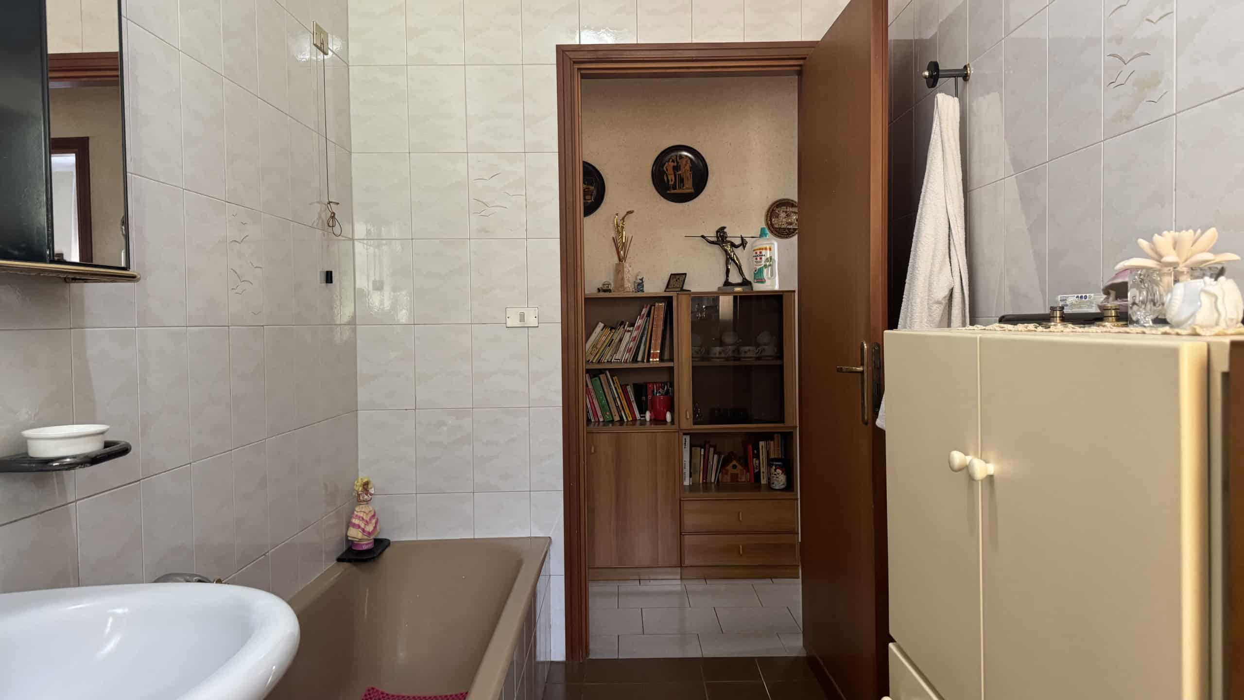 Interno di un bagno con vasca, armadio e pareti rivestite di piastrelle chiare.