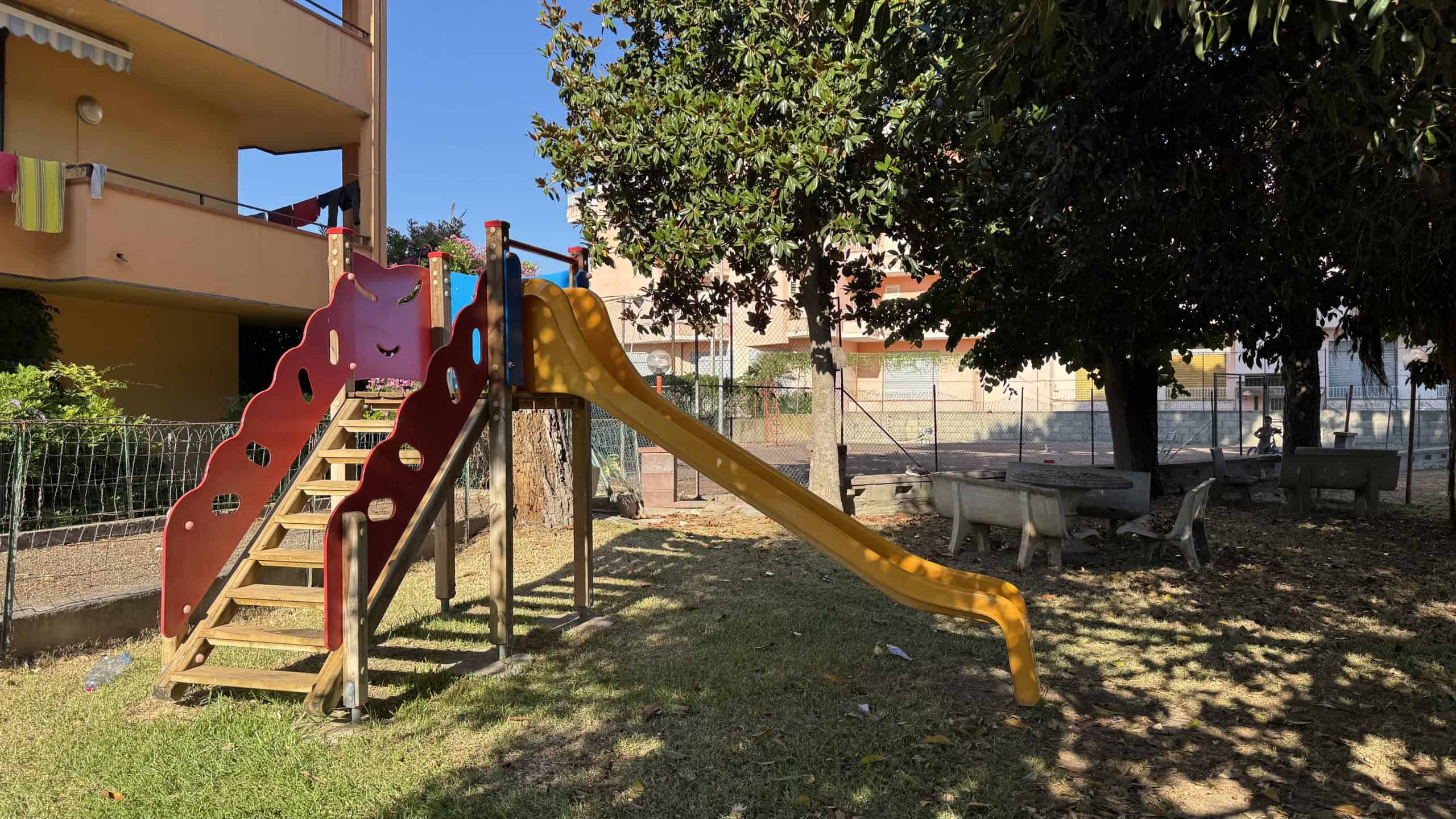 Area giochi per bambini con scivolo e altalena in un cortile condominiale ombreggiato da alberi.
