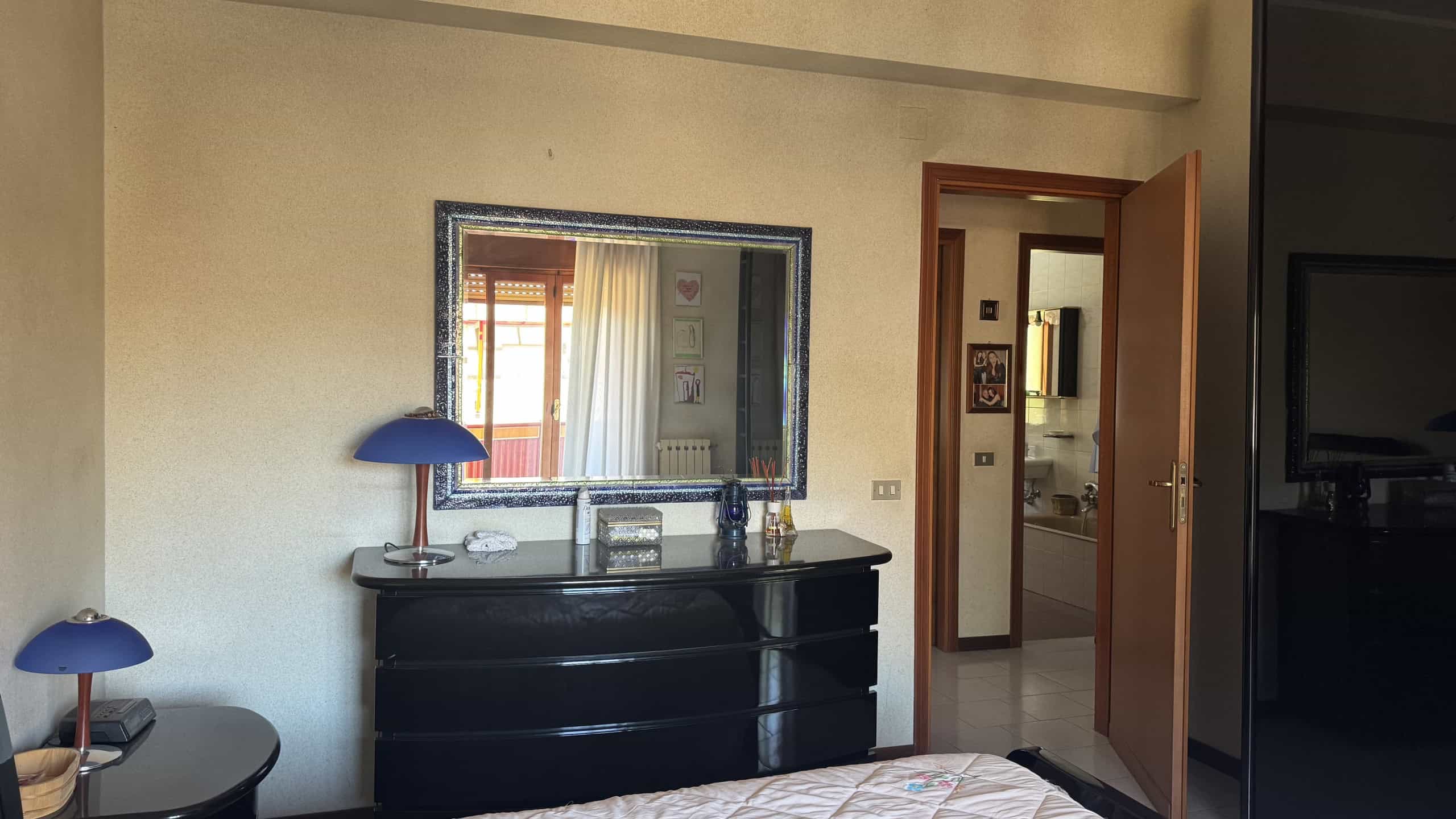 Interno di un appartamento a Messina con mobile nero, specchio e lampade blu, vista sulla porta e sulla stanza adiacente.