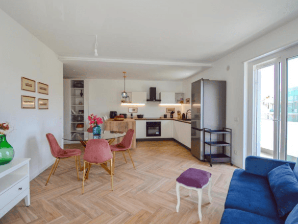 Interno di un appartamento moderno con cucina a vista, zona pranzo e soggiorno con divano blu, arredamento elegante e ampie finestre che lasciano entrare molta luce naturale.