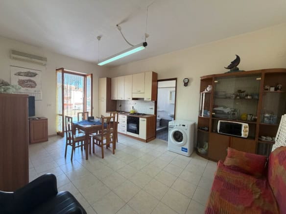 Interno di un appartamento con cucina, zona pranzo e soggiorno, arredato con mobili in legno e divano rosso, finestra con vista esterna.